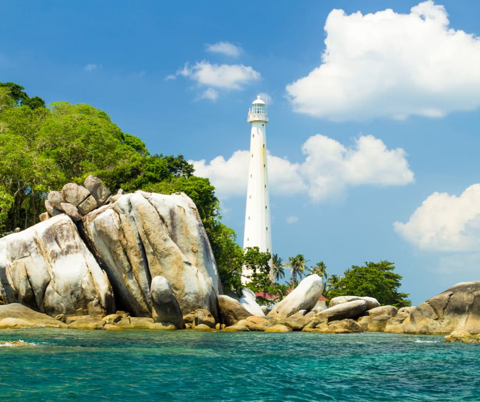 Tall white lighthouse rising behind granite boulders and green trees along a turquoise coastline