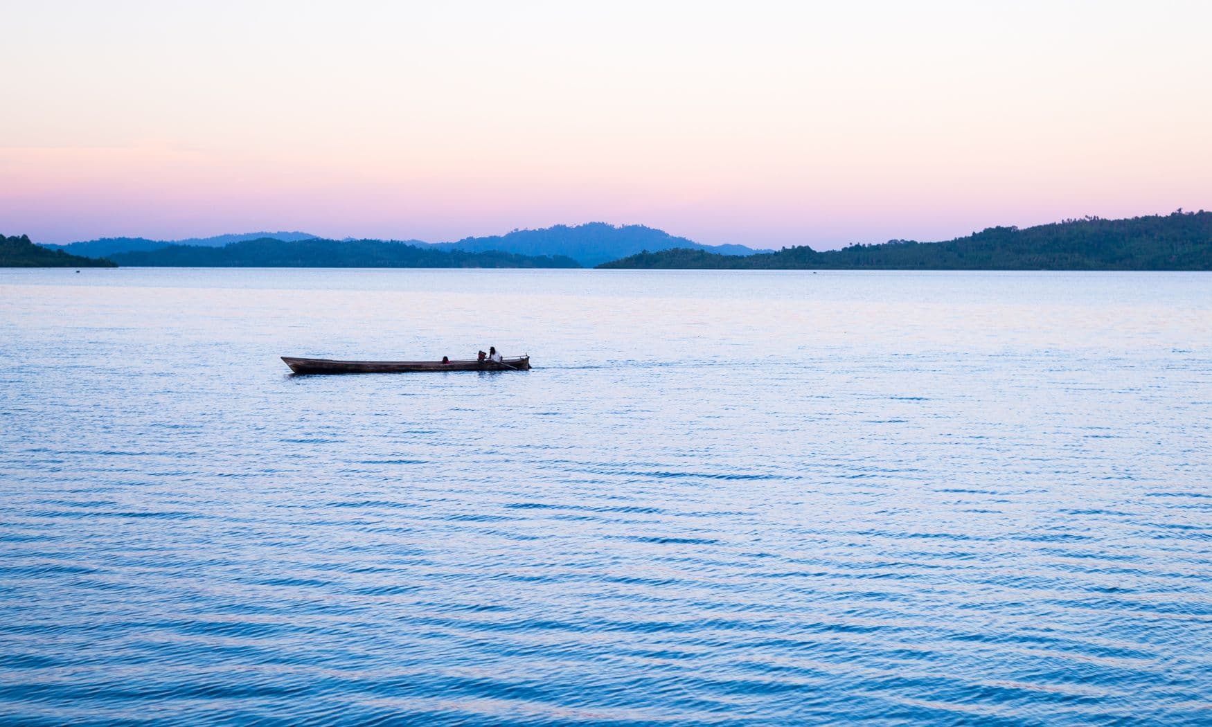 Small wooden boat with two people drifting on calm water at dusk, silhouetted hills along the horizon