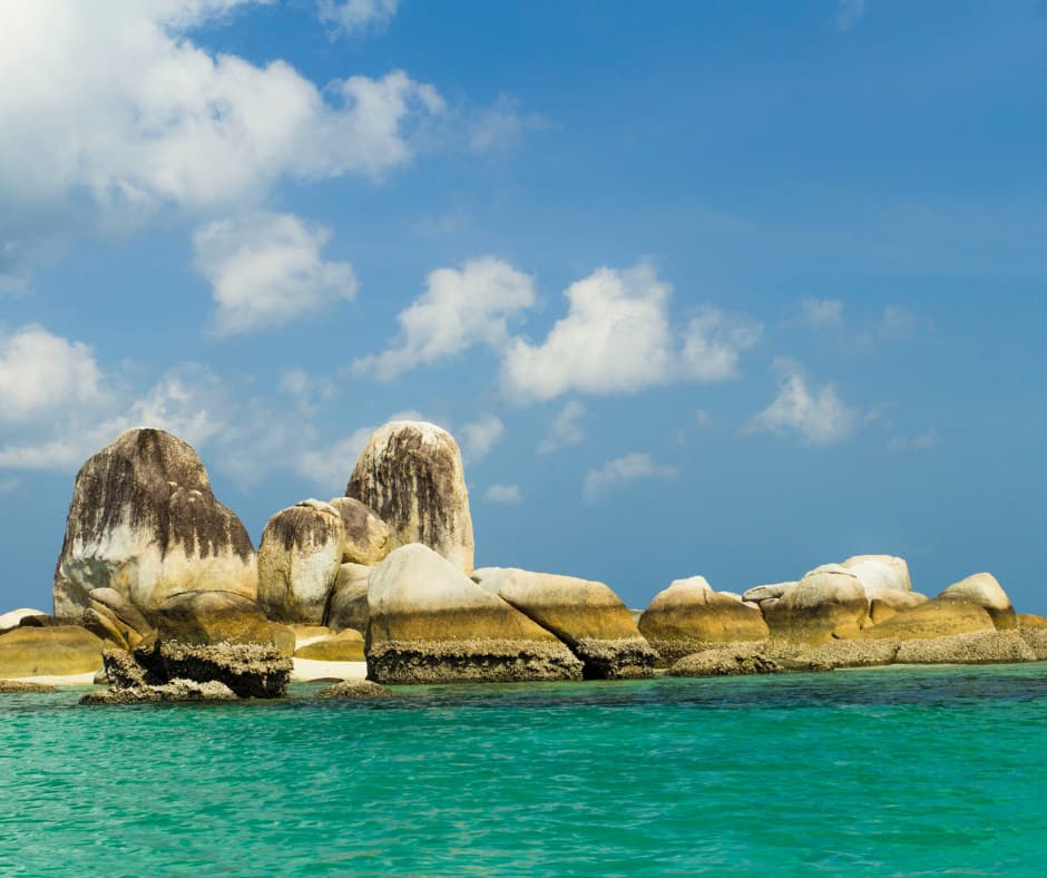 Large weathered granite boulders rising from turquoise sea water under a blue sky with scattered clouds
