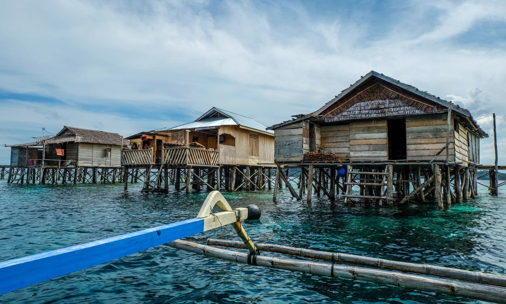 Wooden stilt houses over clear turquoise water, seen from a boat with a blue outrigger in the foreground