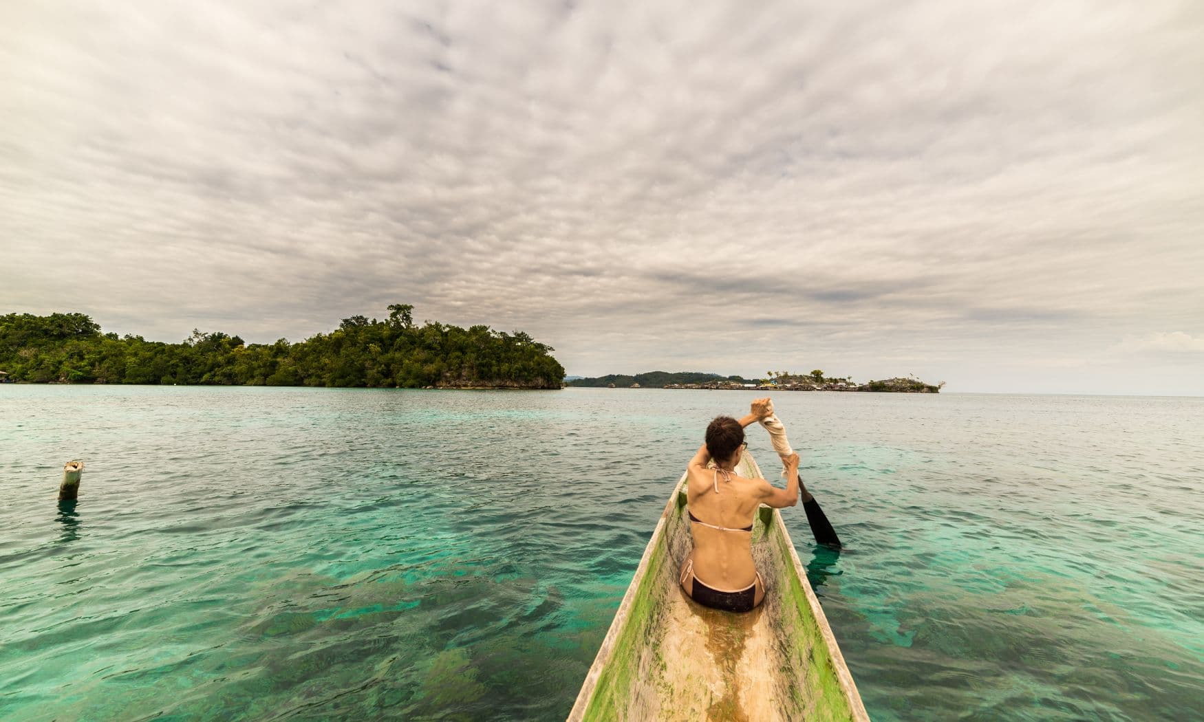 Woman sitting in a wooden canoe photographing the sea, with forested islands visible under an overcast sky