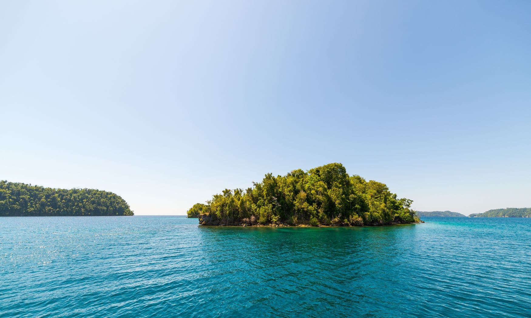 Small forested island rising from turquoise water with a larger tree-covered island visible on the horizon
