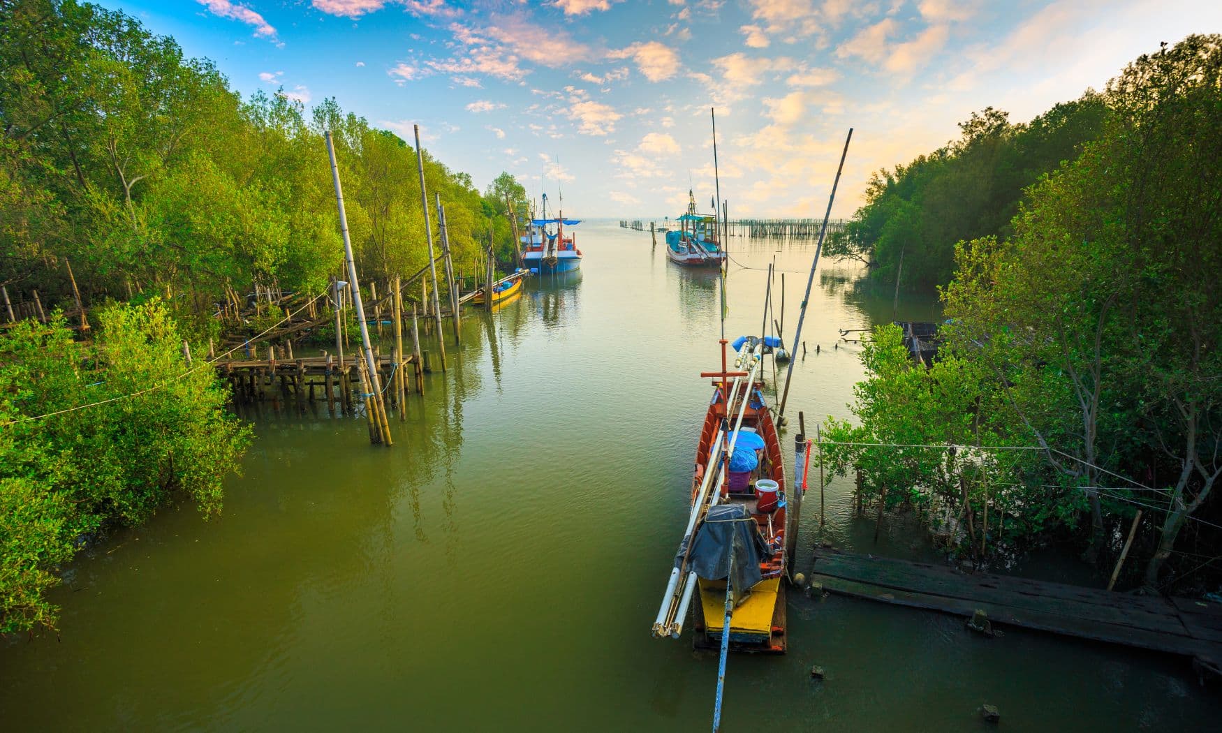 Wooden fishing boats moored in a calm mangrove-lined channel with wooden jetties under a colorful sky