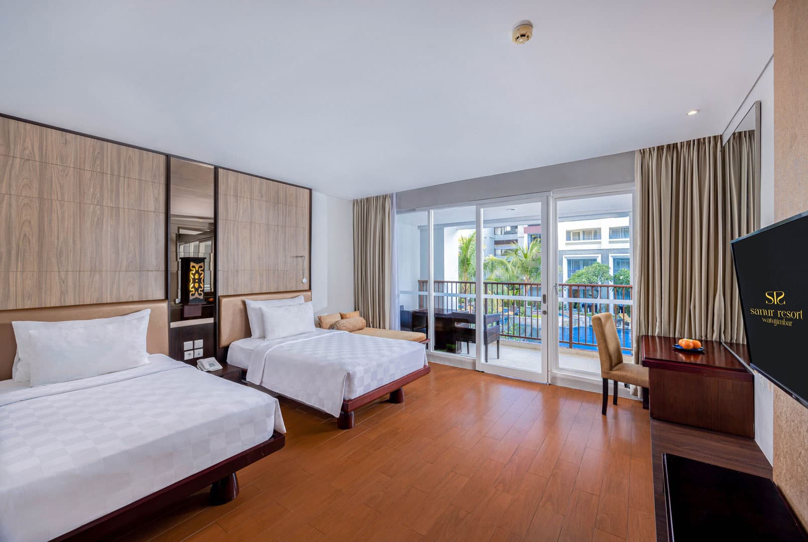 Twin beds with white linens, wooden floor, TV, balcony doors with curtains, and bedside furnishings in a hotel room at Sanur Resort Watujimbar