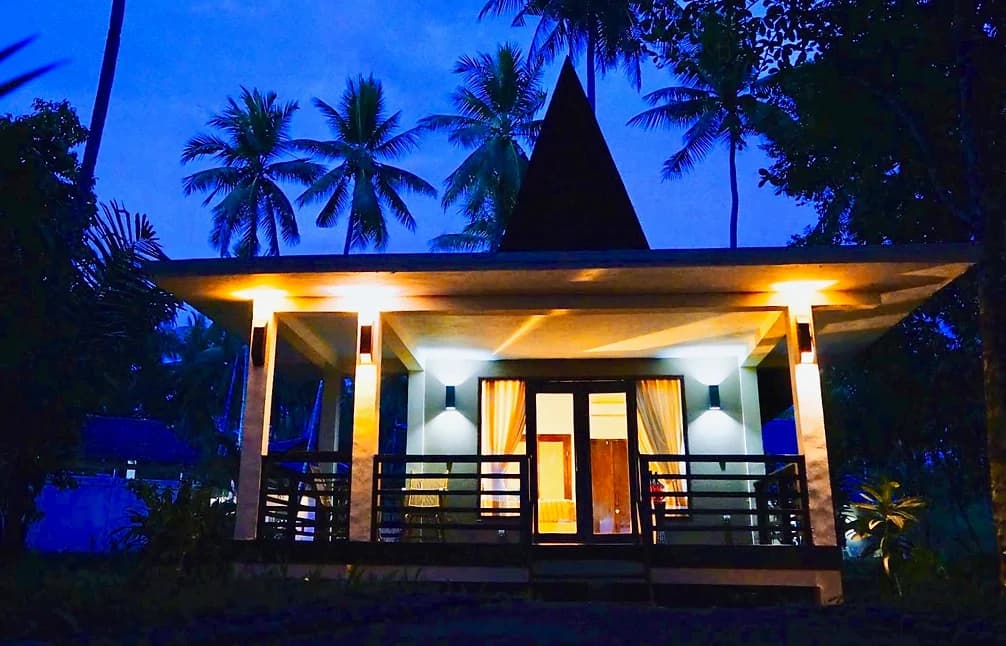 Spice Island Divers bungalow illuminated at dusk with a peaked roof, wooden railings, and palm trees surrounding the structure against a blue twilight sky