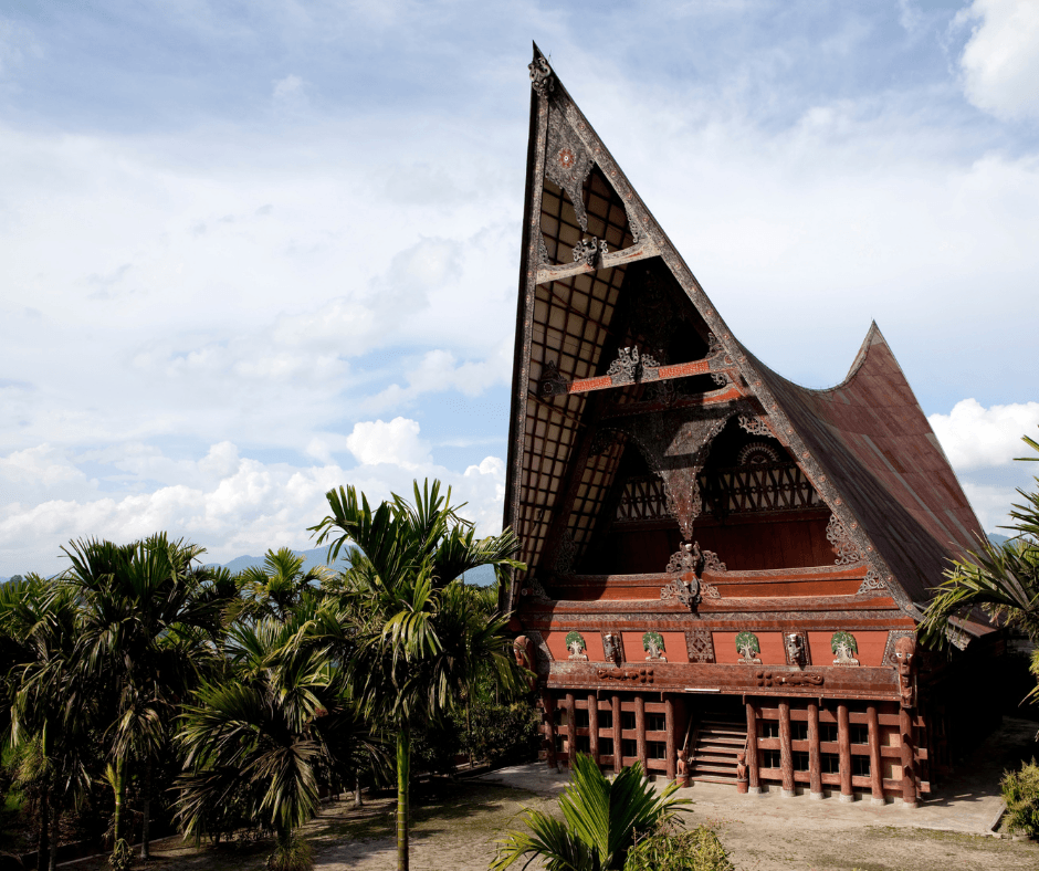 Traditional wooden building with a tall curved saddle-shaped roof and carved decorative panels, surrounded by palm trees