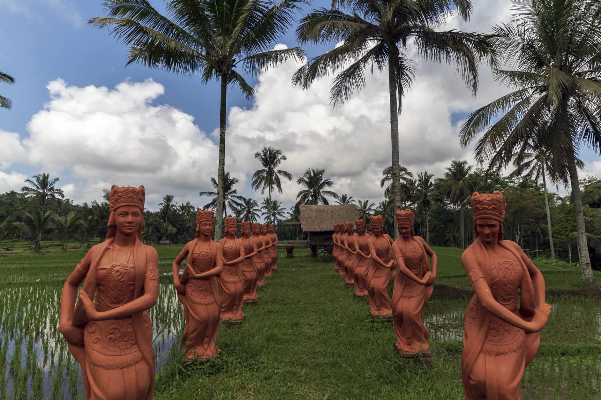 Terracotta statues of figures in traditional dress arranged in a line across a grassy field with rice paddies, palm trees, and thatched structures at Jiwa Jawa Resort Ijen