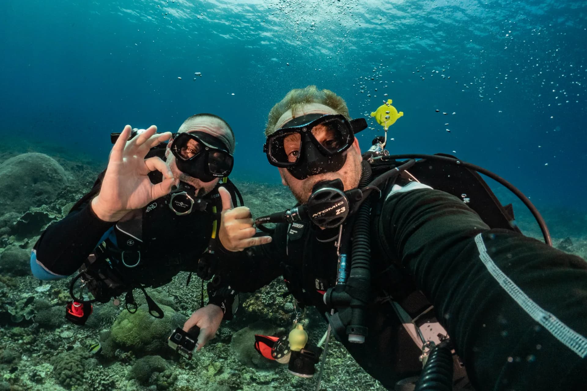 Two scuba divers signaling okay and shaka hand gestures underwater near a coral reef