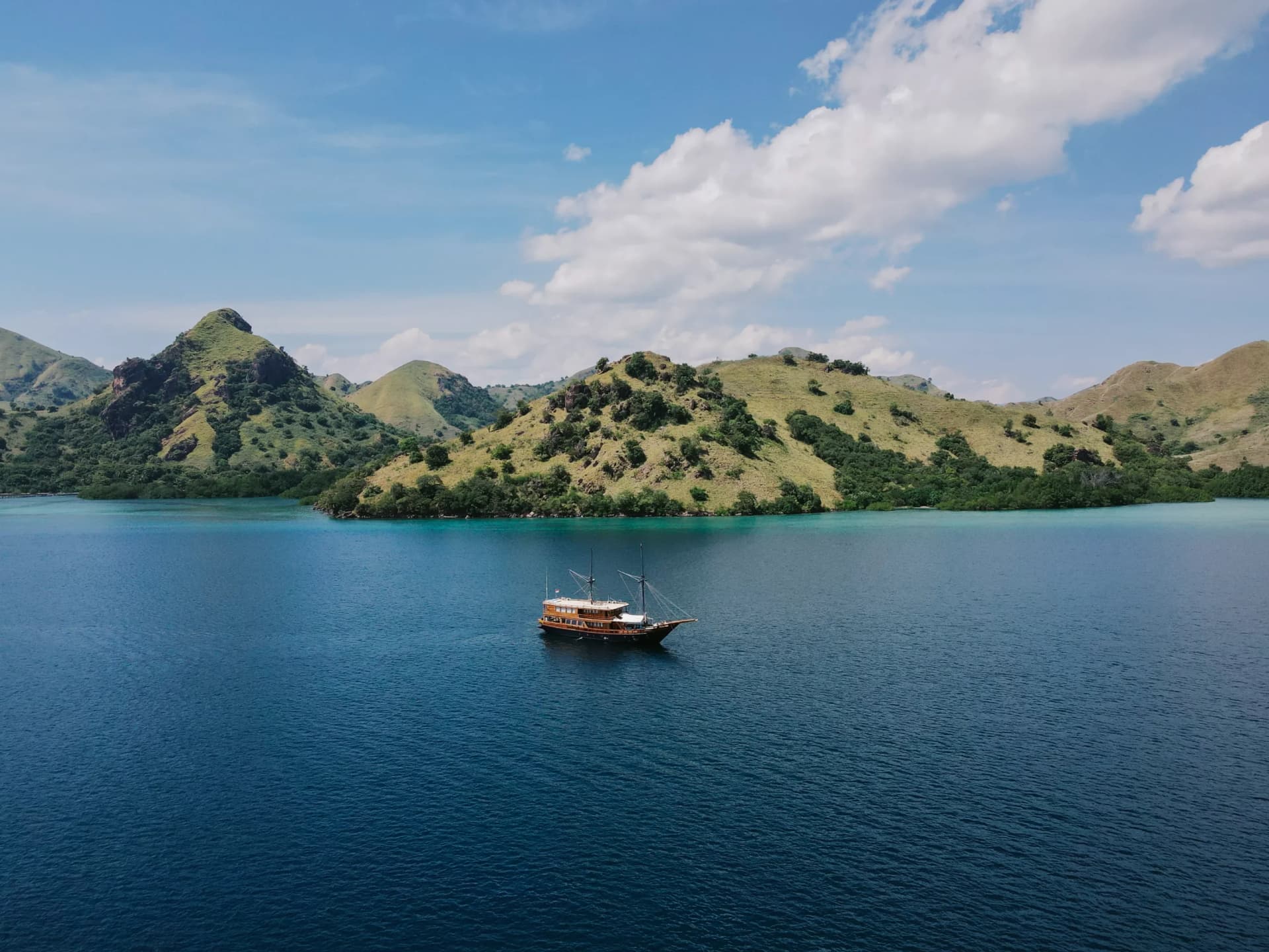 Augustine Phinisi anchored in a calm bay surrounded by green hills under a blue sky with scattered clouds