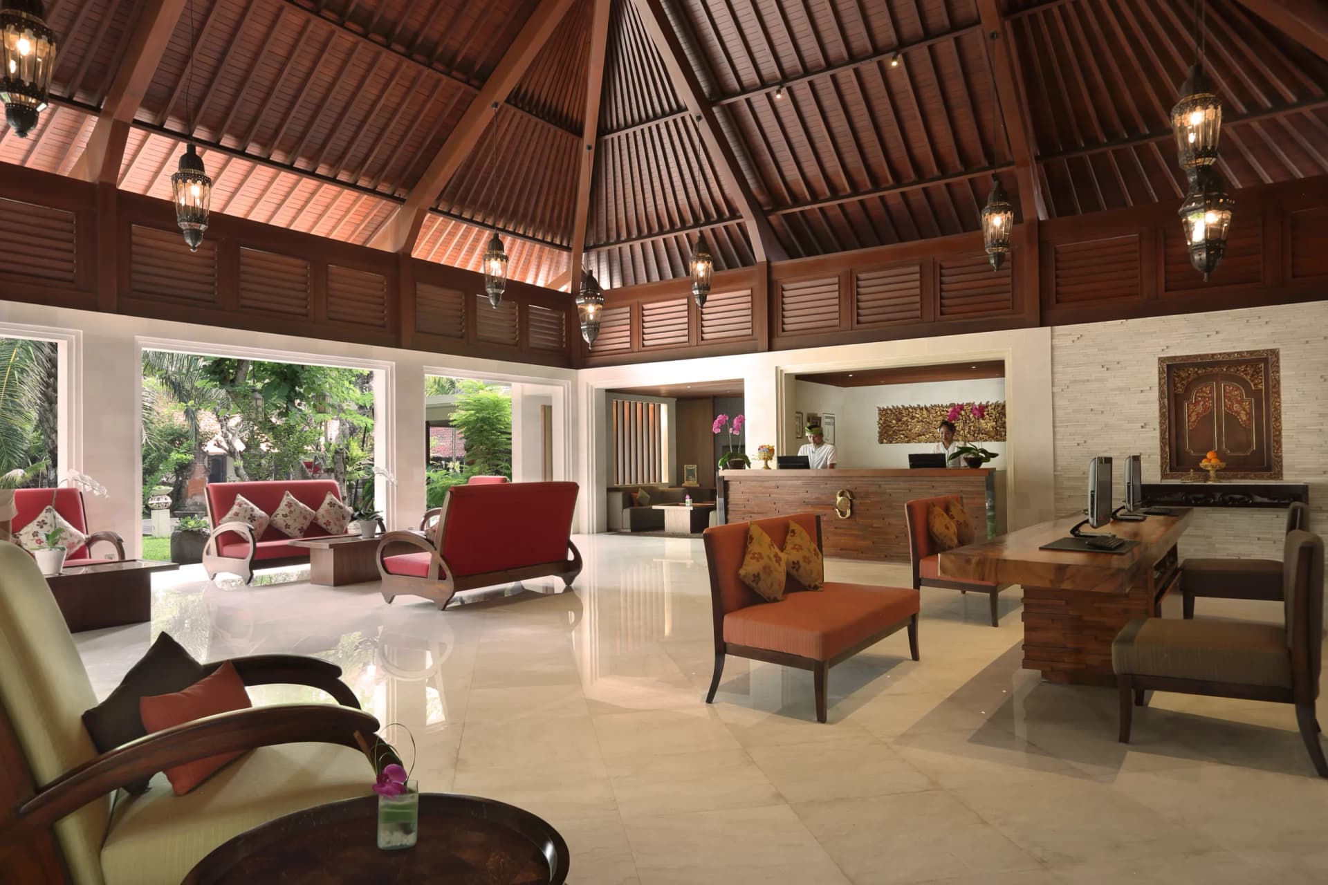 Griya Santrian's open-plan lobby and lounge with wooden ceiling, red upholstered seating, wooden furniture, polished stone flooring, and large windows overlooking gardens