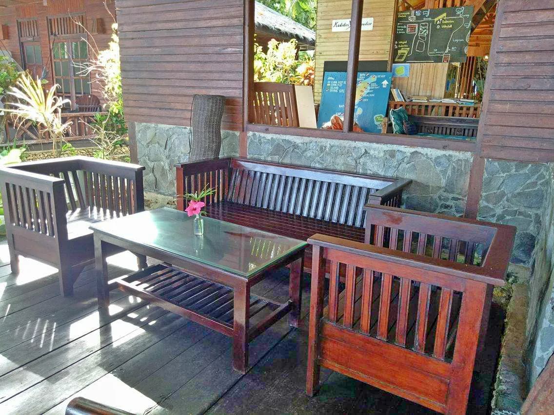 Wooden seating and a table on a covered patio area with stone walls, plants, and a view into an indoor space at Kadidiri Paradise Resort and Dive Center