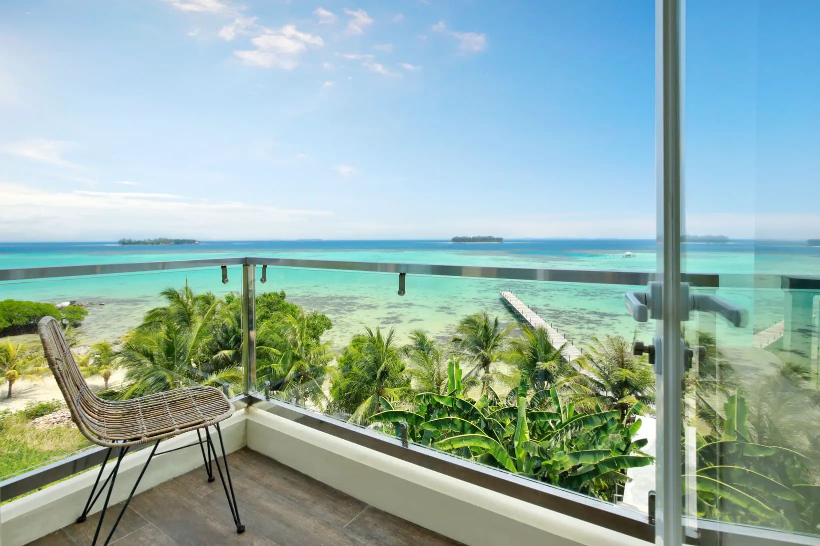 Seating on a balcony with glass railing overlooking tropical vegetation and turquoise water with a distant horizon