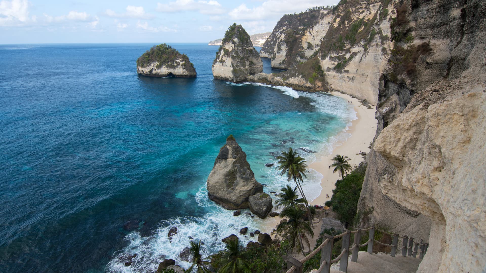 Aerial view of limestone cliffs, rock pillars, and turquoise water bordering a sandy beach with palm trees