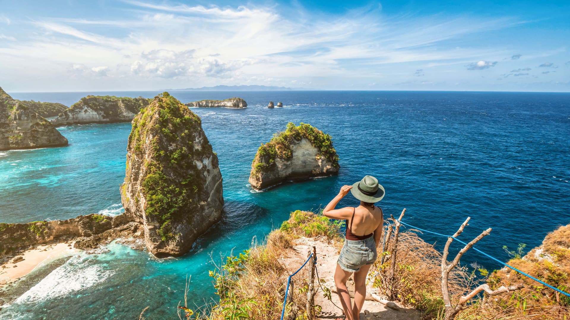Woman in a hat looking out over turquoise water at tall green-topped rock formations rising from the sea