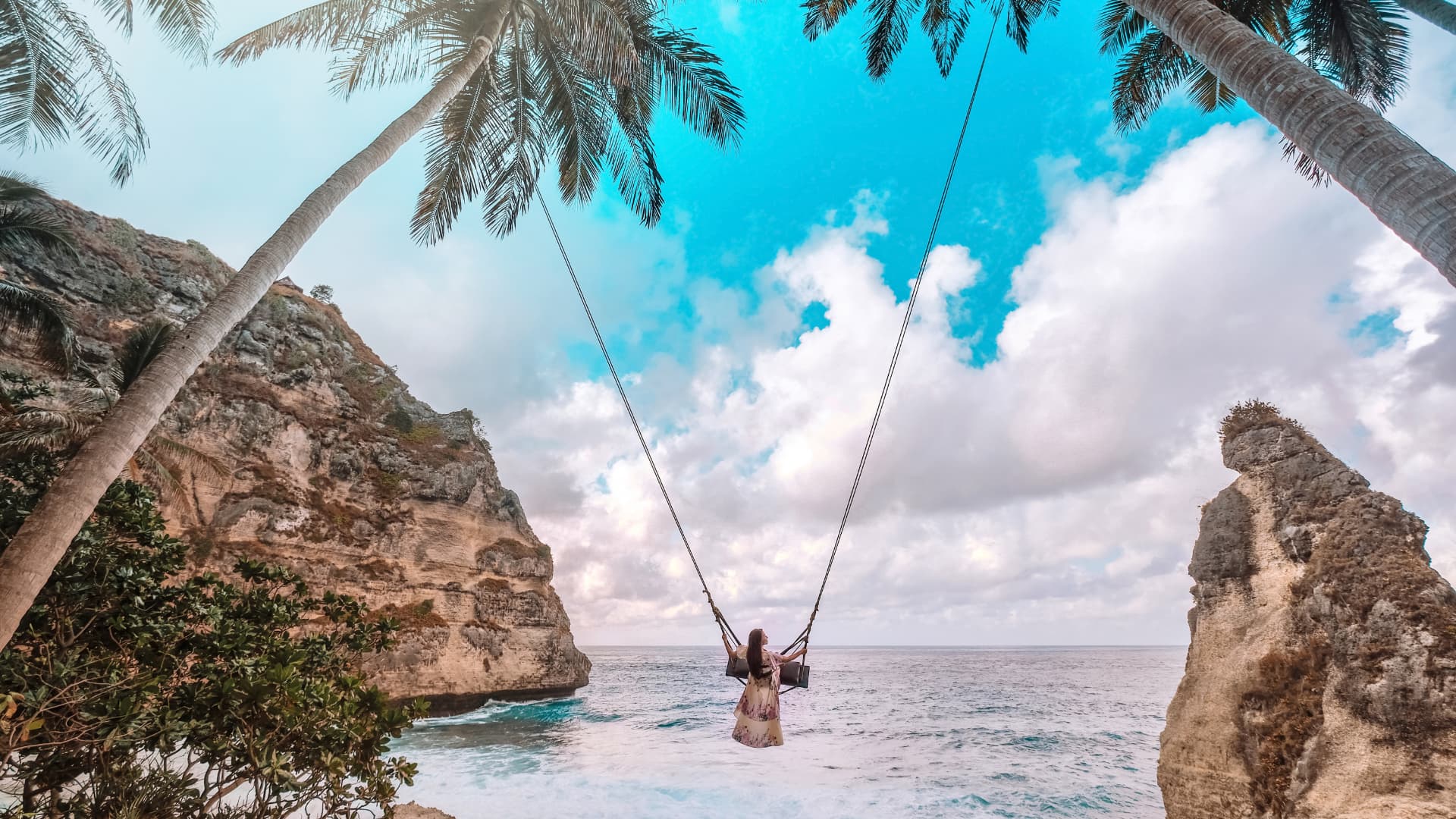 A person on a rope swing suspended between palm trees over the ocean, framed by tall rock formations