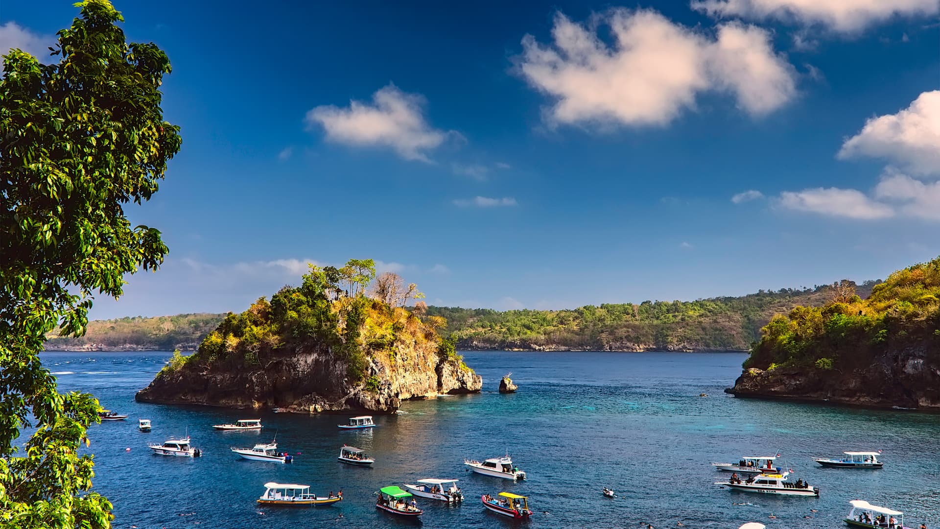 Small boats anchored in a turquoise bay beside a tree-covered rocky outcrop under a blue sky with scattered clouds