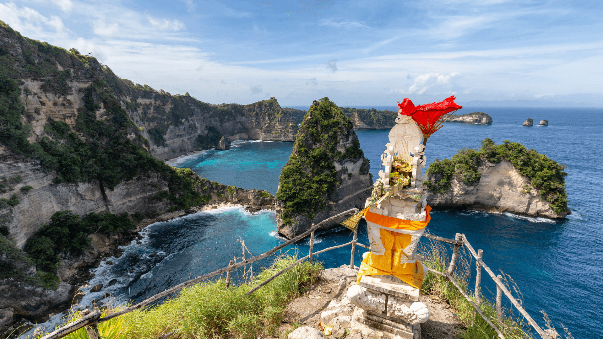 Statue draped in orange and red cloth on a cliff edge overlooking turquoise water, rocky islets, and green headlands