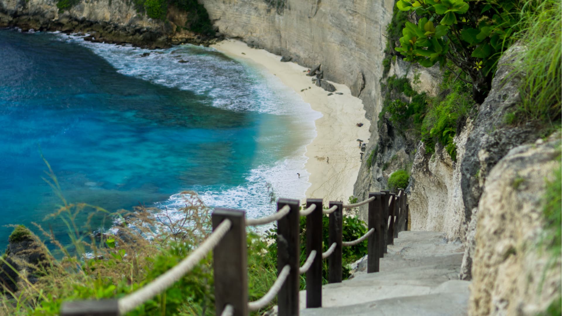 Stone steps with a rope railing descend a cliffside toward a curved white sand beach and turquoise water