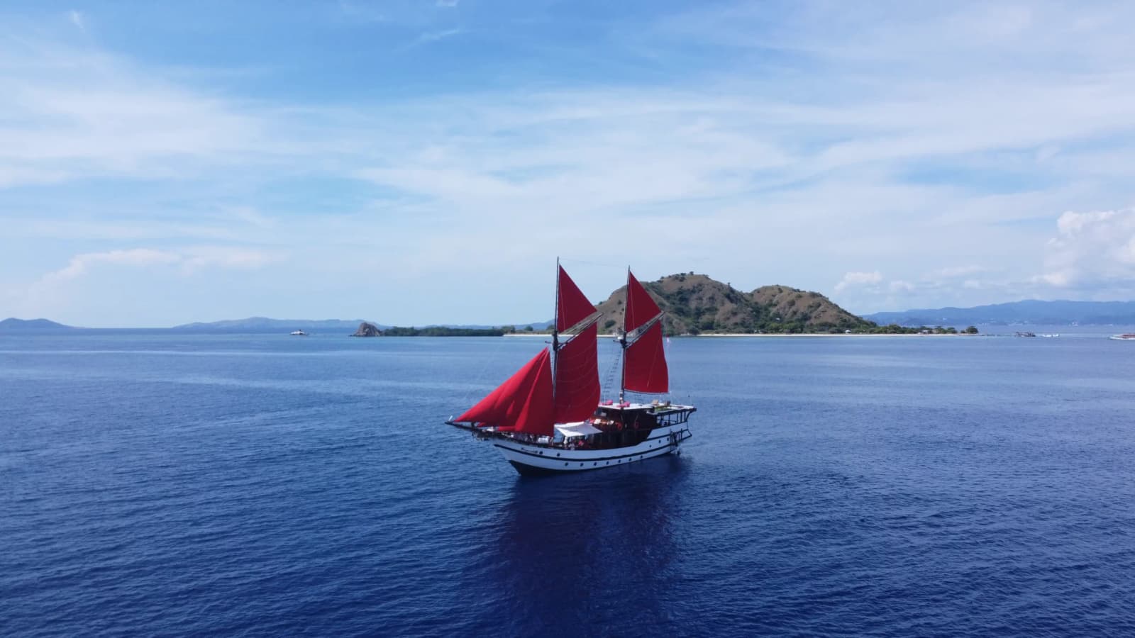 Aerial view of the SuprabA traditional sailing vessel with red sails on calm blue water near a hilly coastline