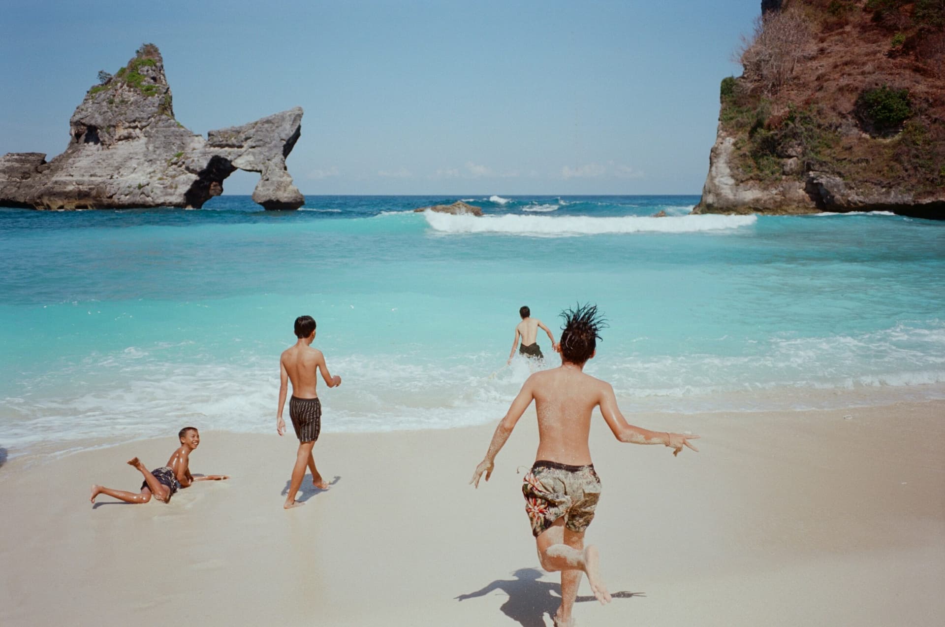 Four people running and playing in shallow water on a white sand beach with rock formations offshore
