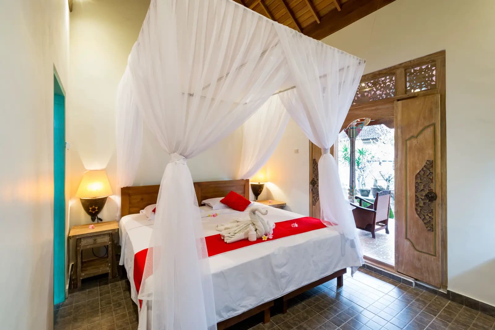 Bedroom at Umasari Rice Terrace Villa with four-poster bed draped in white curtains, red cushions, bedside lamps, wooden doors and carved panels opening to a terrace