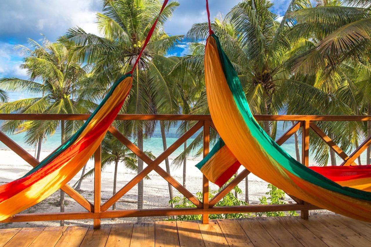 Colorful fabric hammocks hang from a wooden deck railing with palm trees and water visible beyond