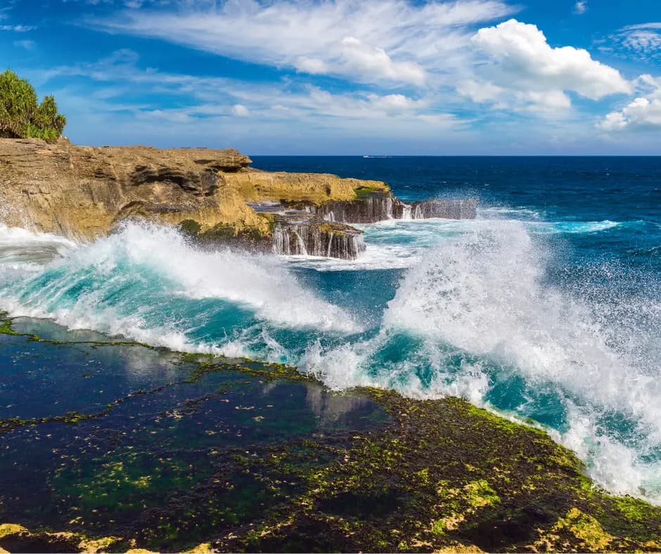 Waves crashing against a rocky, moss-covered coastline beneath a limestone cliff with a small waterfall