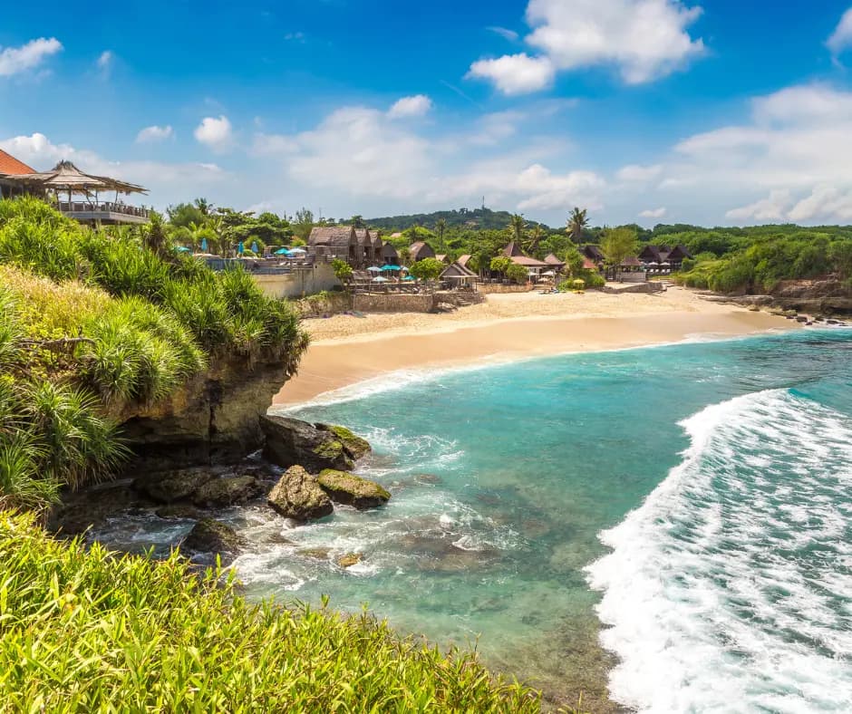 Cliffside view over a curved sandy beach with turquoise water, waves, and thatched-roof buildings among trees