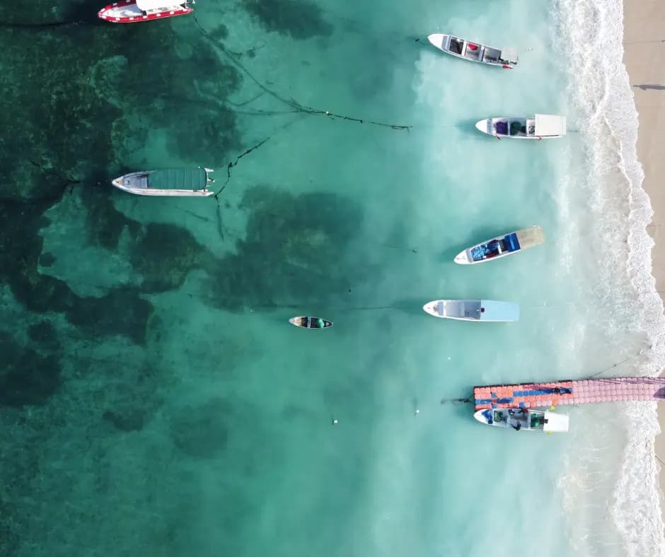 Aerial view of small boats anchored along a shallow reef edge near a sandy shoreline with breaking waves