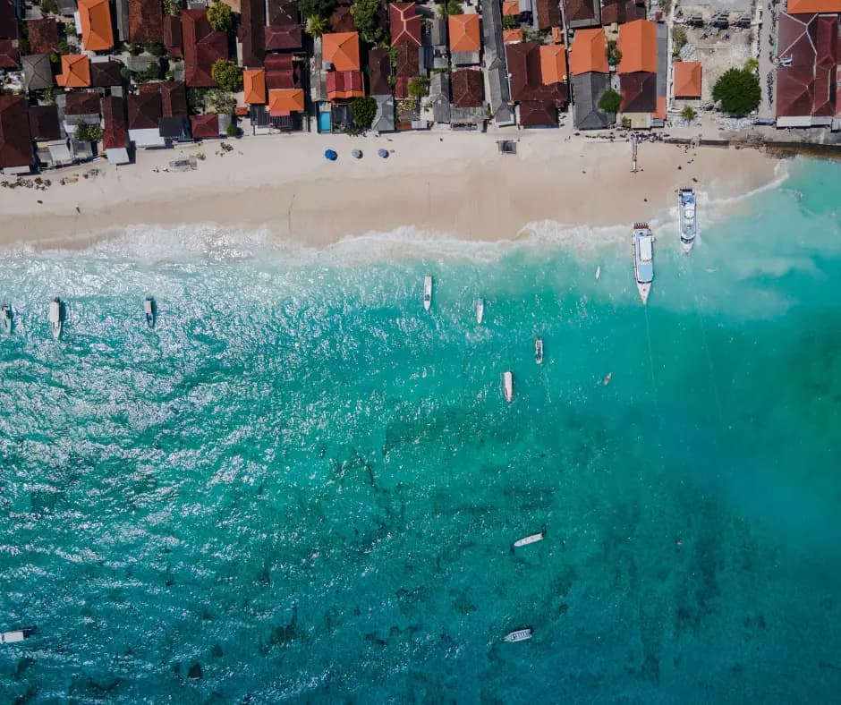 Aerial view of a beachfront lined with red-roofed buildings, boats anchored in turquoise water and waves along the shore