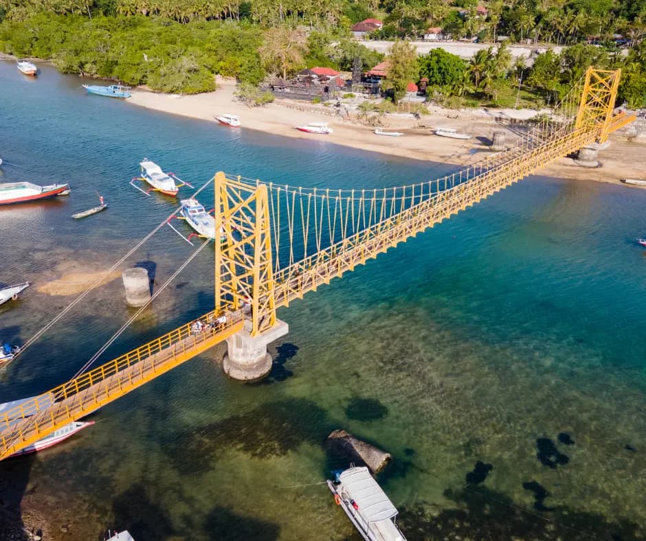 Aerial view of a yellow suspension bridge crossing turquoise water between a beach and moored boats