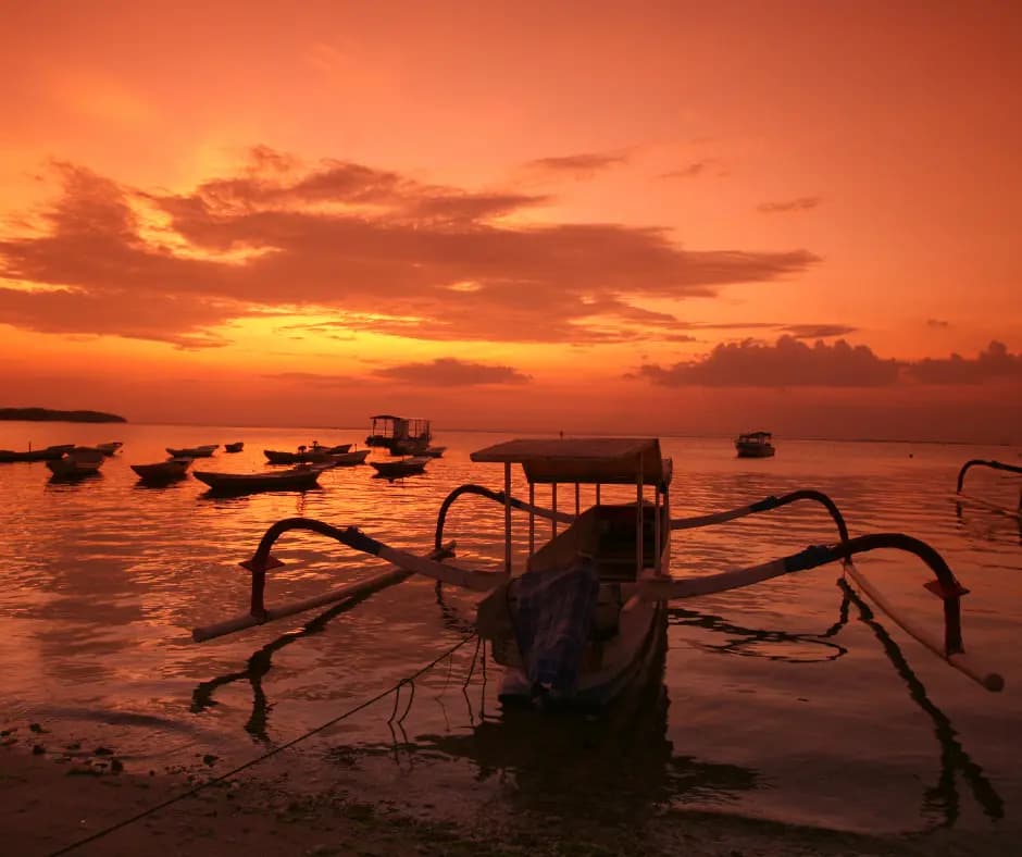 Traditional outrigger fishing boats silhouetted on calm water under an orange sunset sky