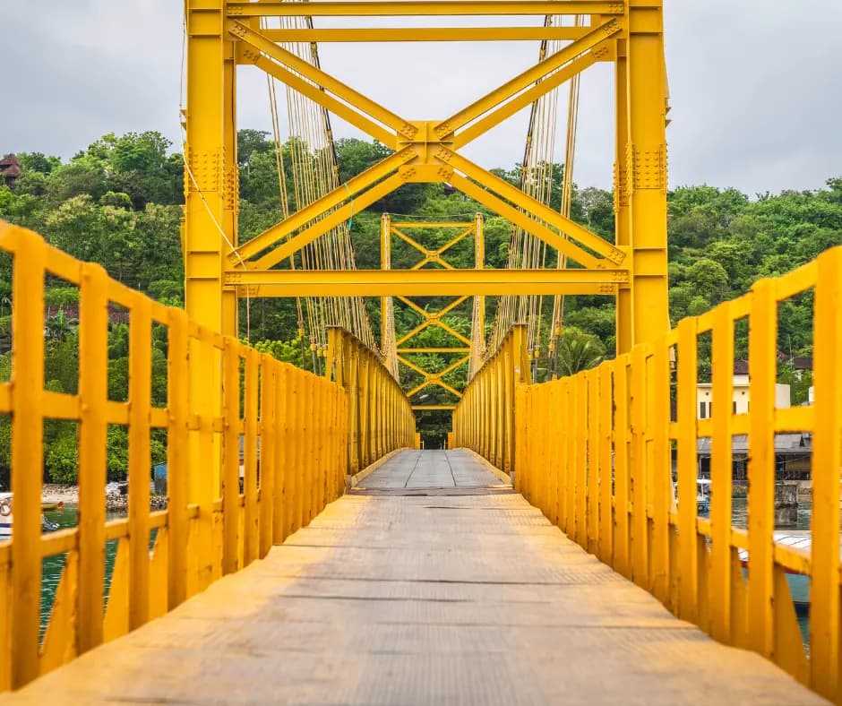 Yellow wooden footbridge with matching railings leading to a yellow-framed suspension tower, surrounded by trees