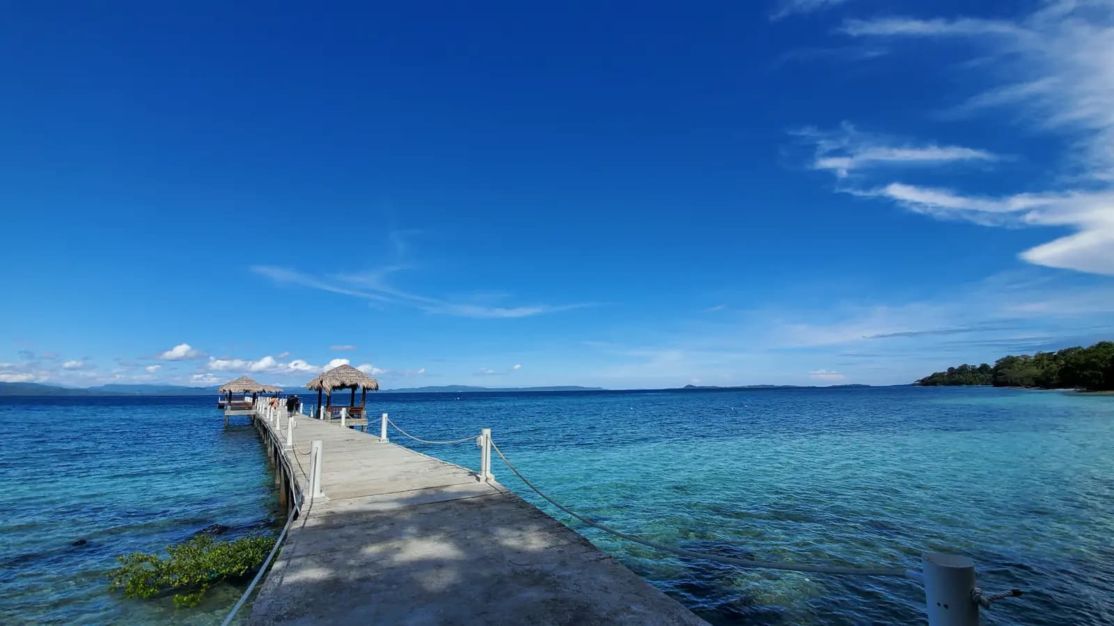 A wooden pier extends into clear blue water with thatched-roof structures at the end, under a bright sky
