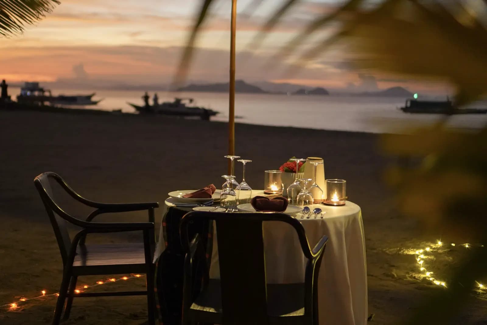 A candlelit dinner table on a beach at dusk with glassware, place settings, and a view of boats and water in the background