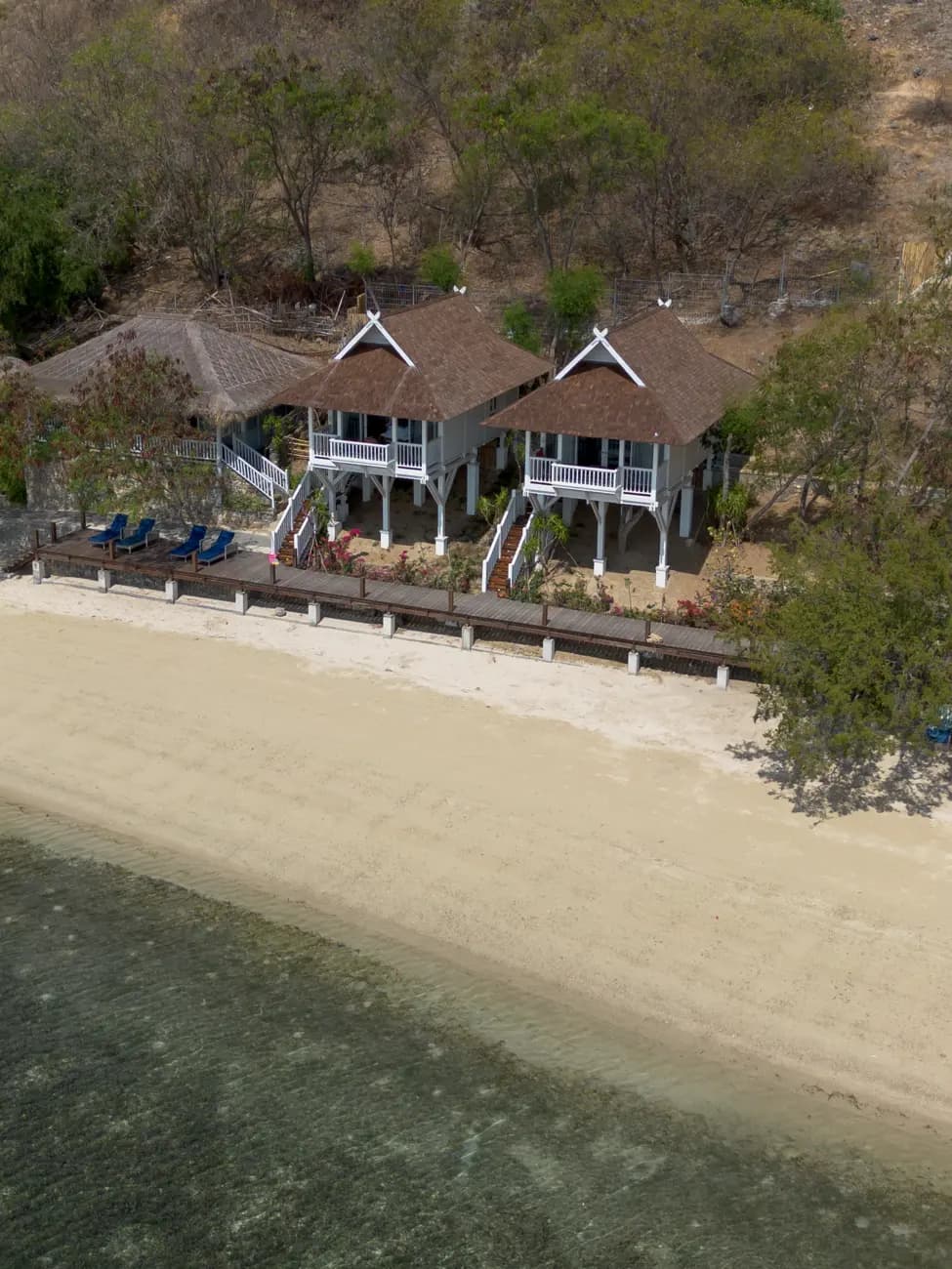 Aerial view of two residential buildings with white railings and pitched roofs beside a sandy beach with seating and wooden barriers