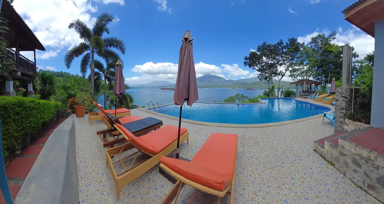 Poolside seating with orange cushions and umbrellas at Bastianos Froggies Lembeh Dive Resort, with infinity pool and water view beyond