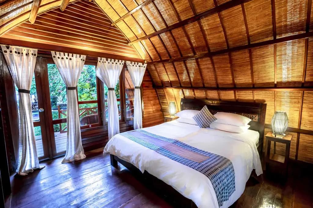 A wooden bedroom with a bed dressed in white linens and a blue patterned runner, wooden ceiling beams, open doors with curtains, and a bedside table with a lamp