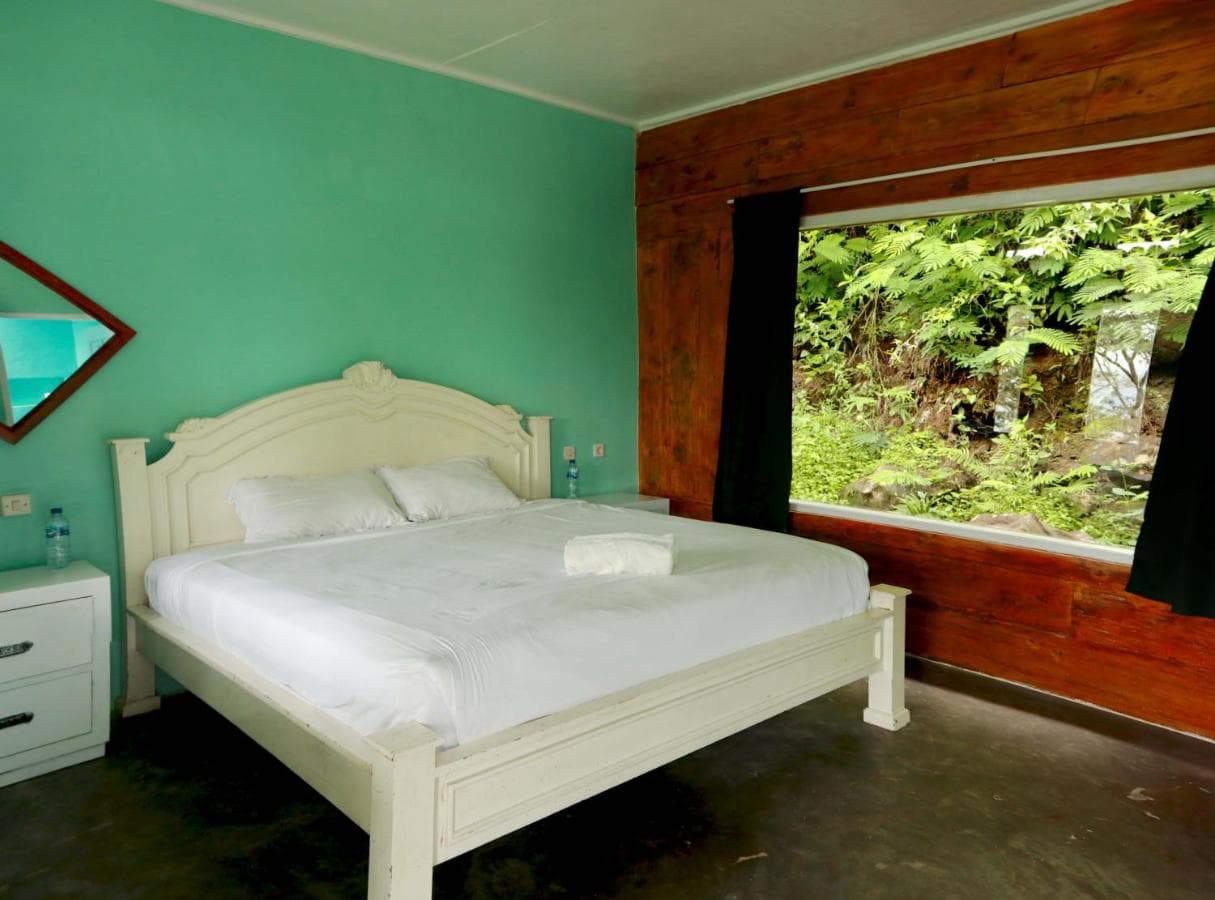 Bedroom with cream-colored bed, turquoise walls, white bedside table, large window with black curtains overlooking greenery, and wooden frame