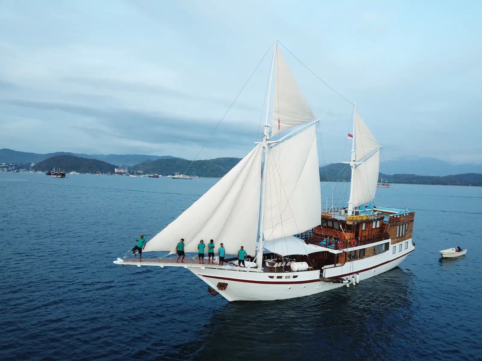 Lady Grace liveaboard under full white sails on calm water with crew standing on its bow platform and hills in the distance