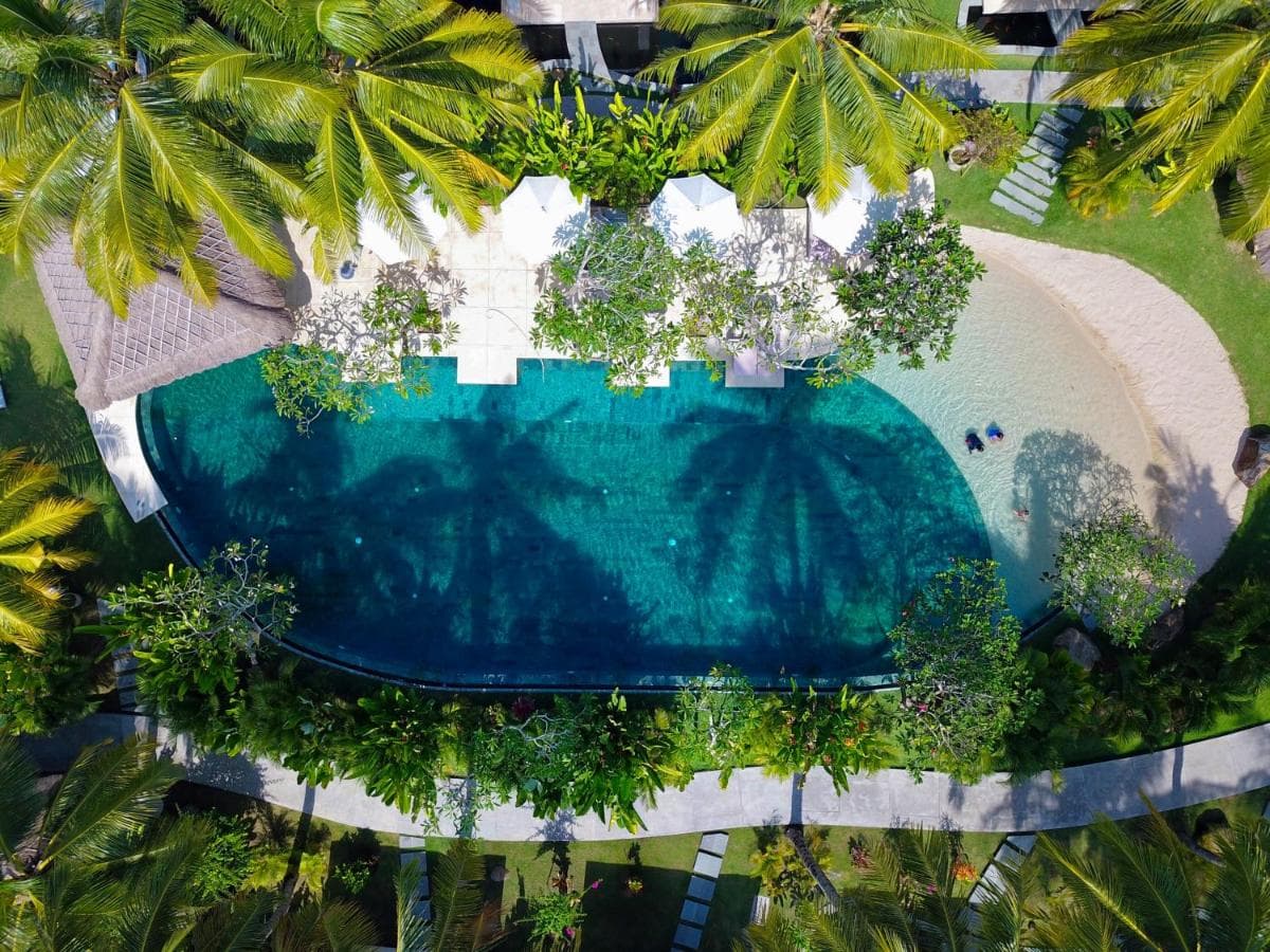 Aerial view of Jivana Resort's pool surrounded by palm trees, white structures, sandy beach, and manicured gardens
