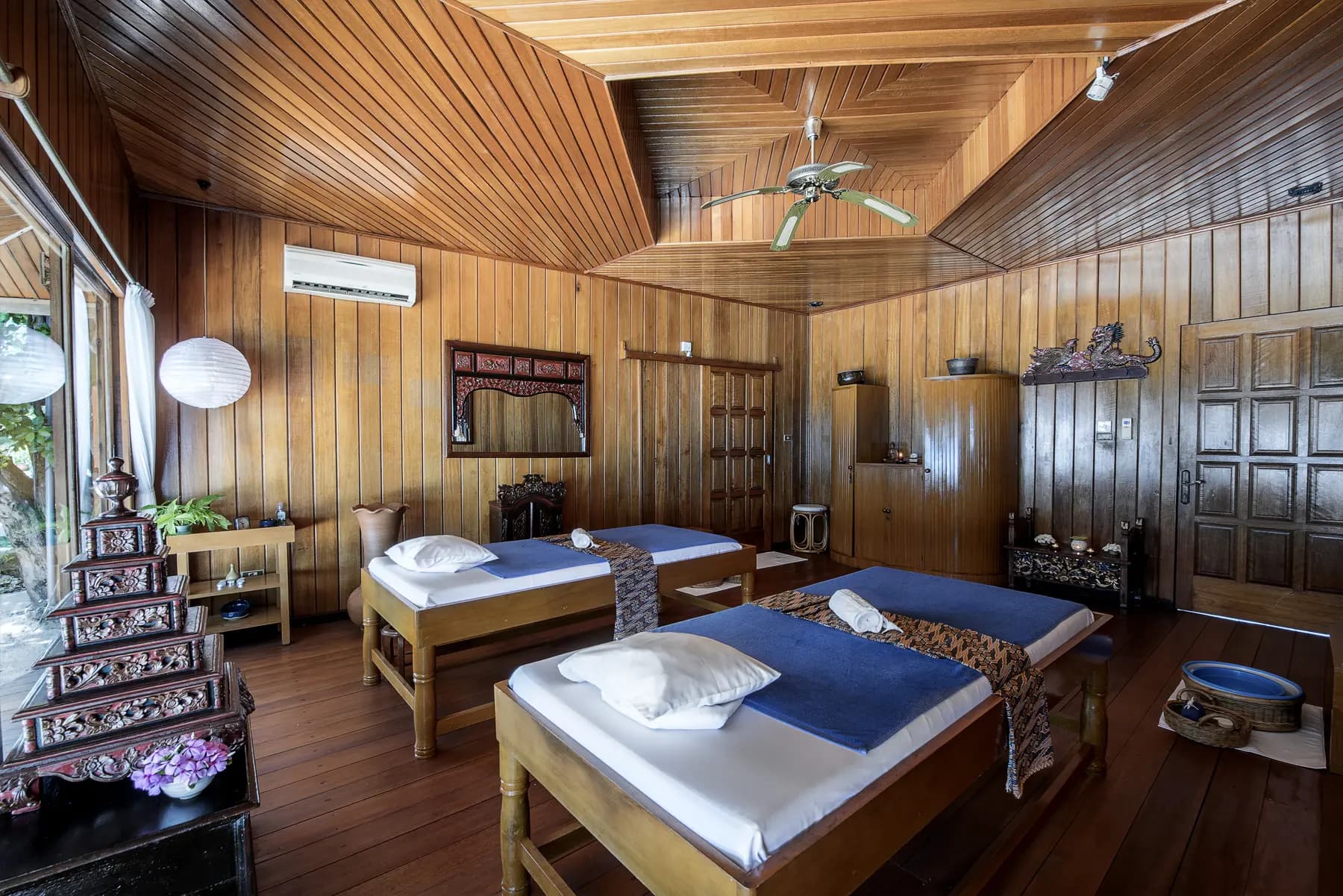 Wooden bedroom with two beds with blue mattresses and white pillows, wooden furniture, ceiling fan, and carved wooden decorative elements