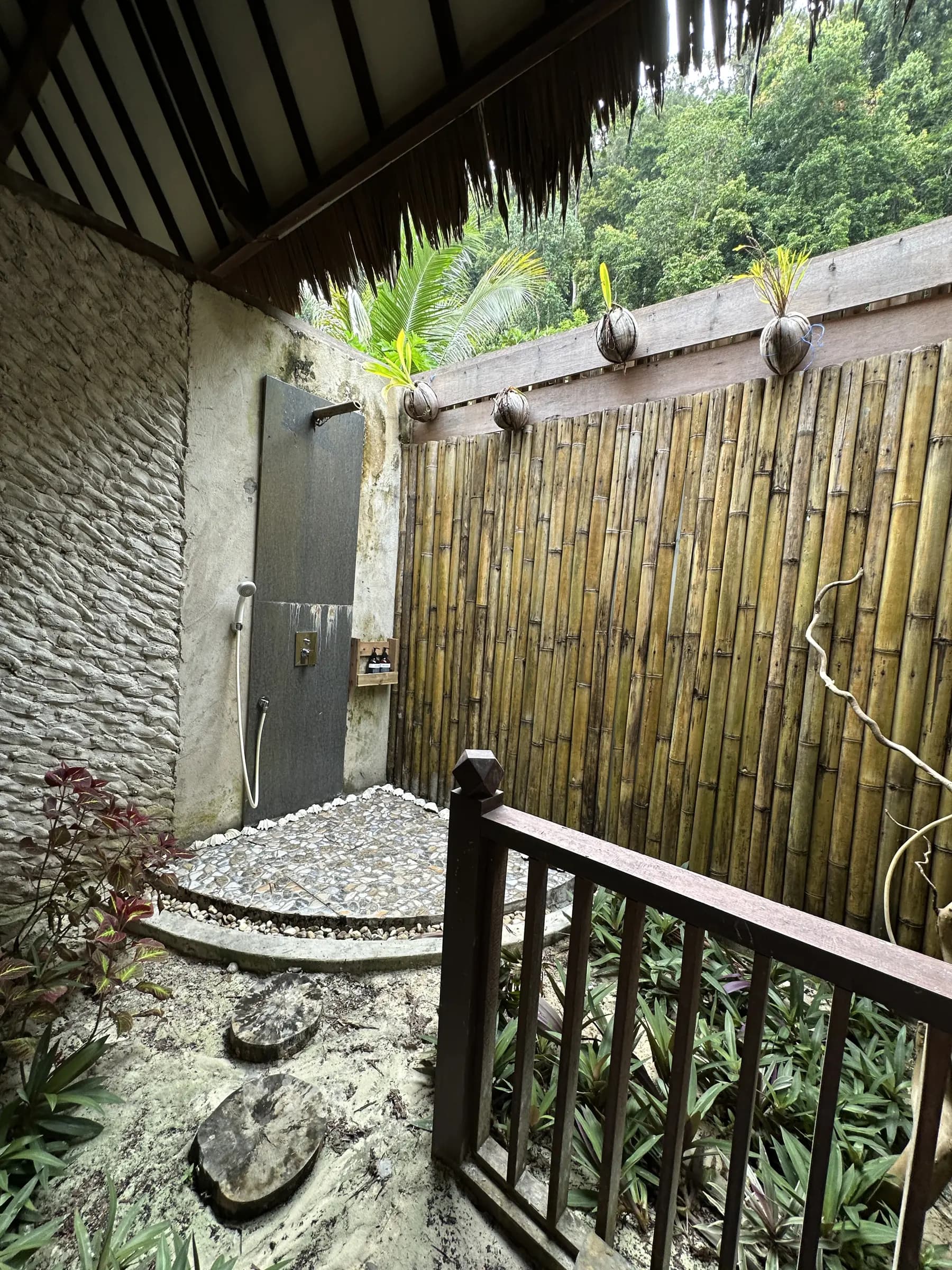 Outdoor shower area at Yenatar Resort Raja Ampat with stone walls, bamboo screening, palm plants, and stone stepping stones on gravel ground