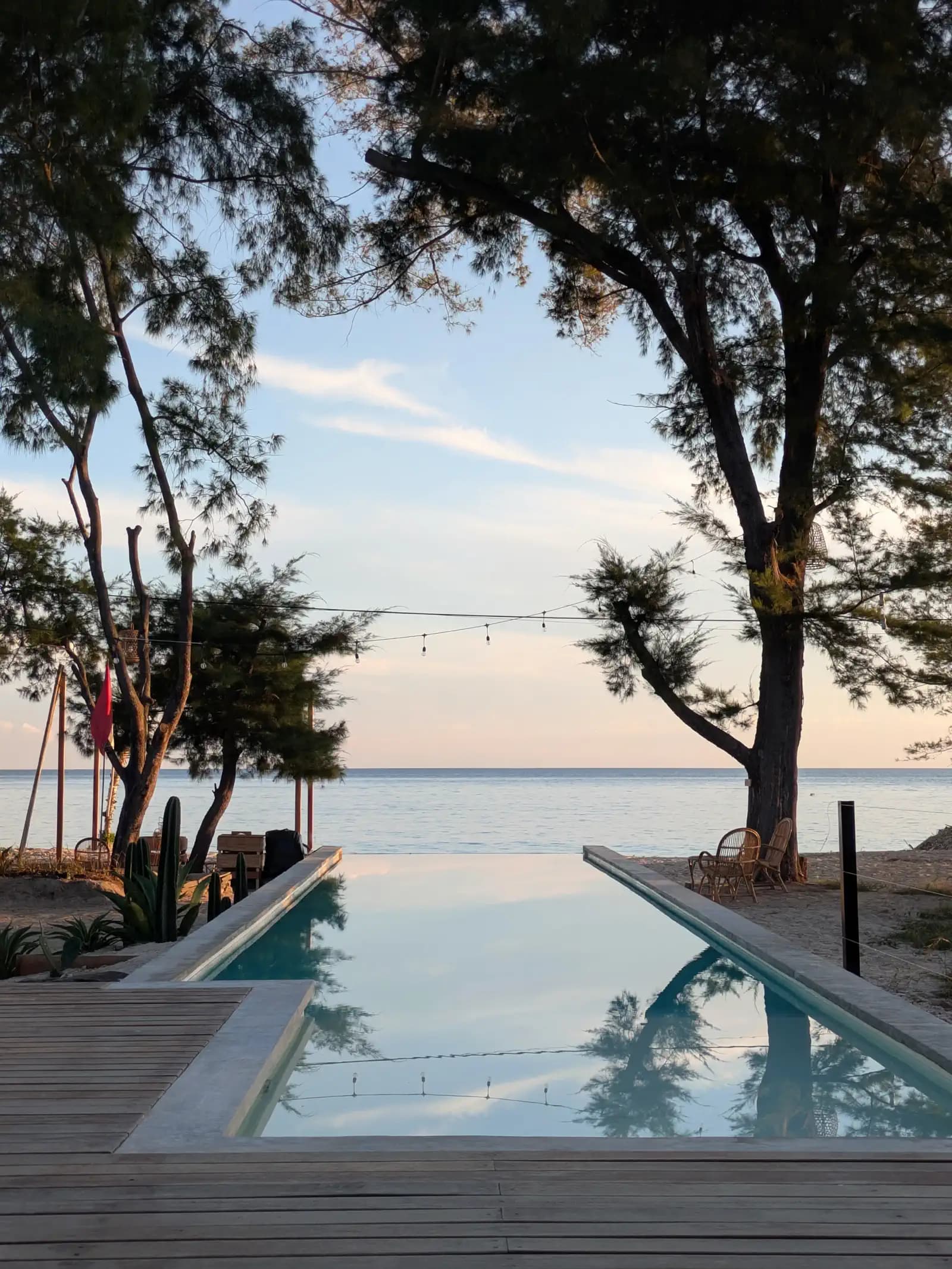 The Beach House Meno's infinity pool reflects trees and sky, extending toward a waterfront view with wooden decking and tropical vegetation on either side