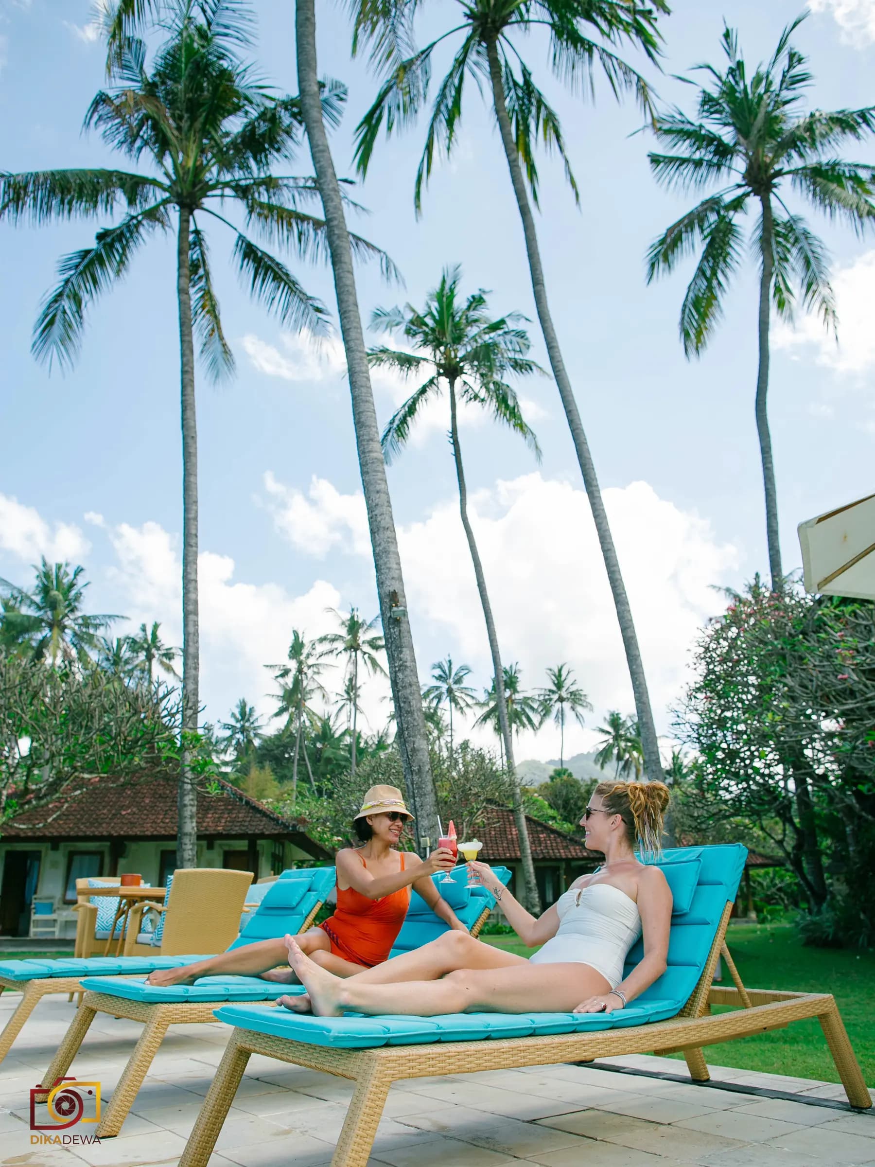 Two guests relax on blue loungers on a lawn at Lotus Bungalows, surrounded by tall palm trees and bungalows