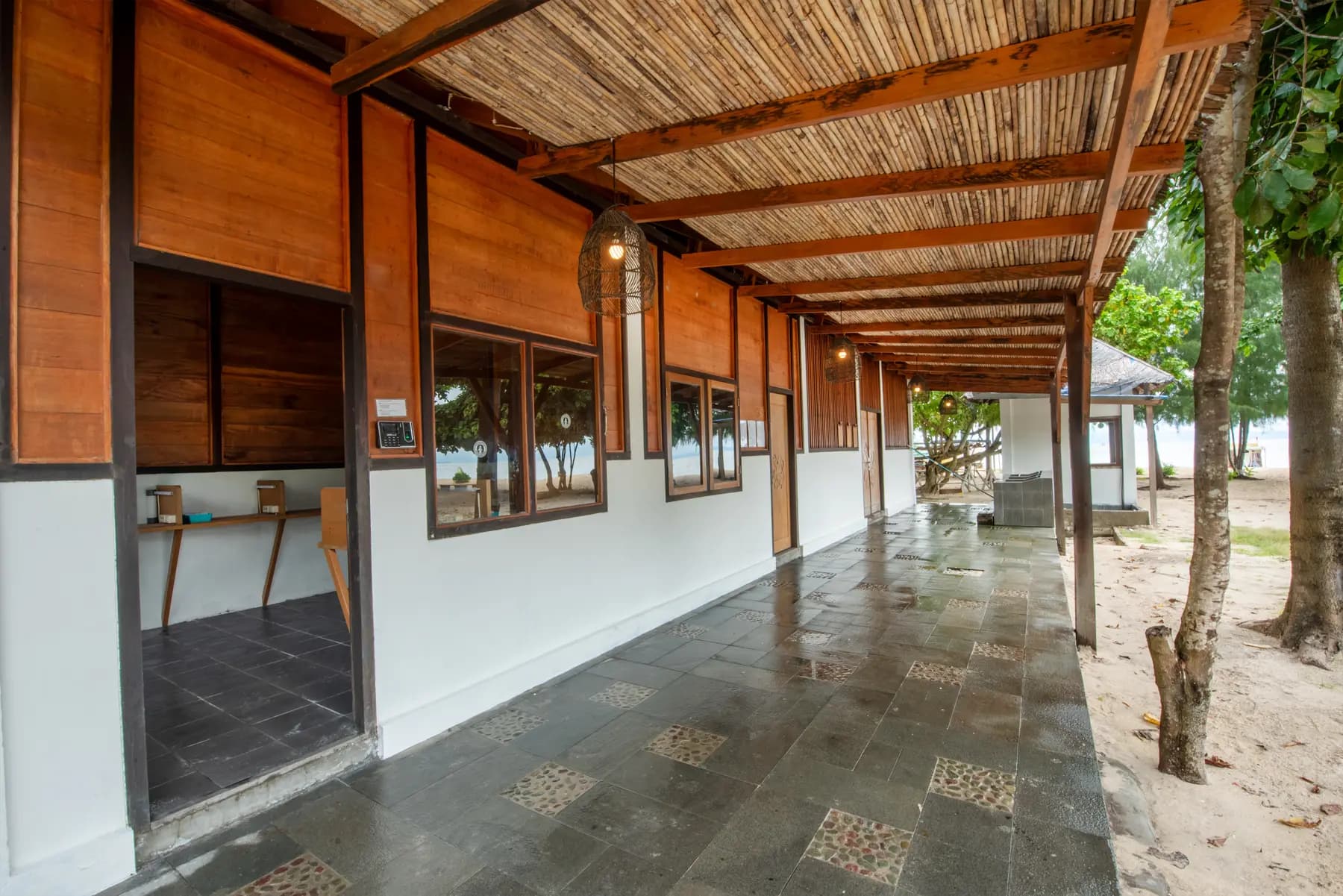 Covered walkway with wooden beams and corrugated roof, white walls, dark tiled floor, wooden doors and windows, trees alongside
