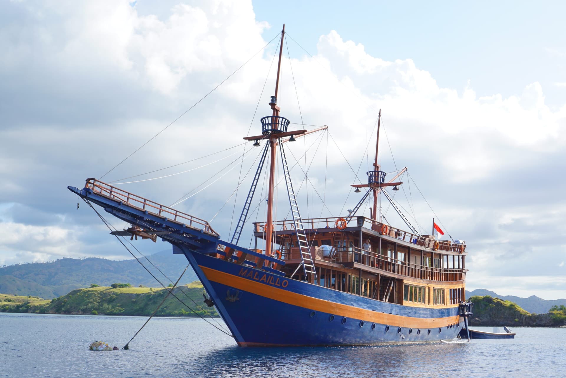 The Malaillo liveaboard boat anchored near a coastline, with two masts and a small tender boat alongside