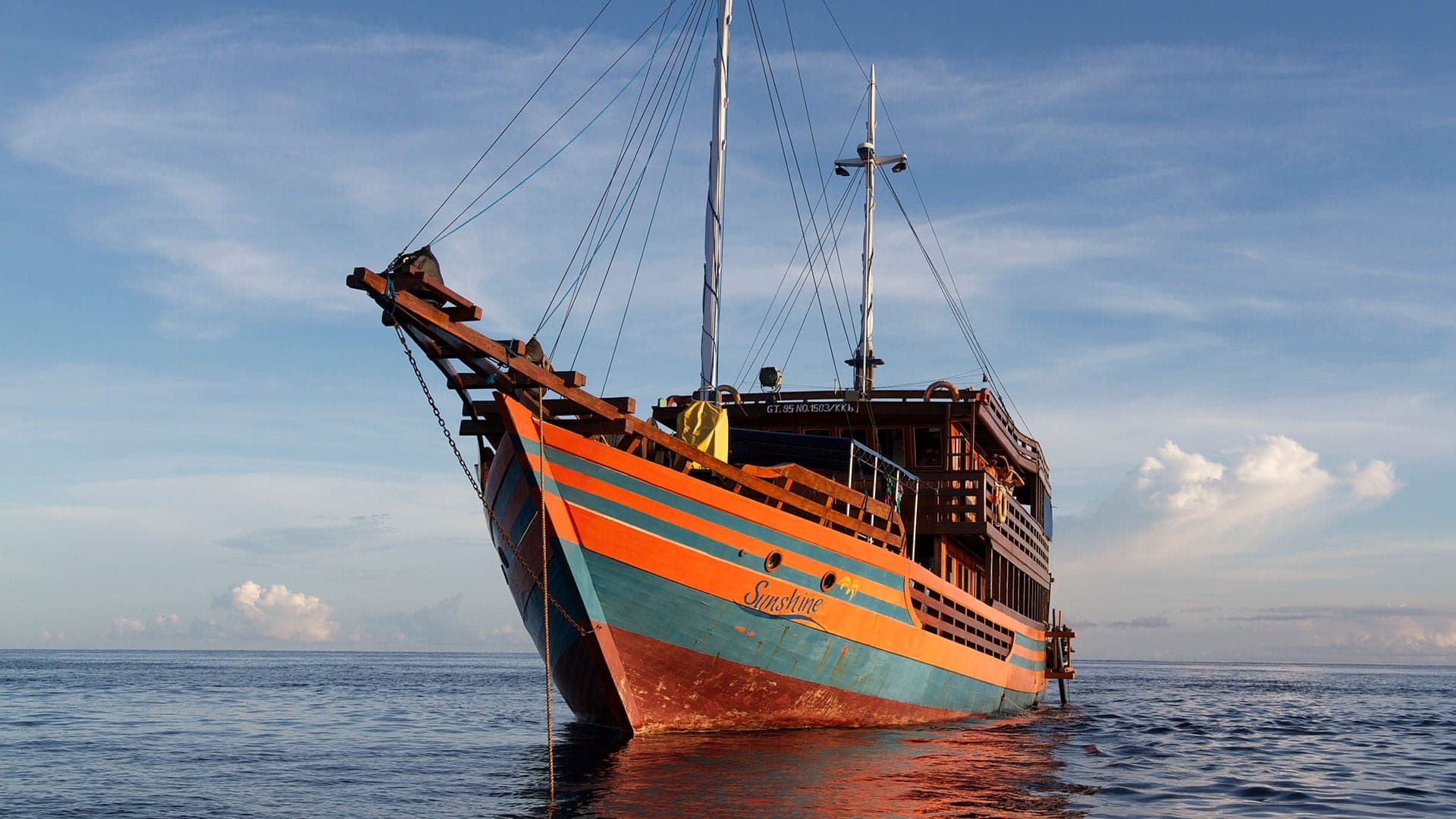 Sunshine Liveaboard wooden boat with orange and teal hull stripes anchored at sea under a partly cloudy sky