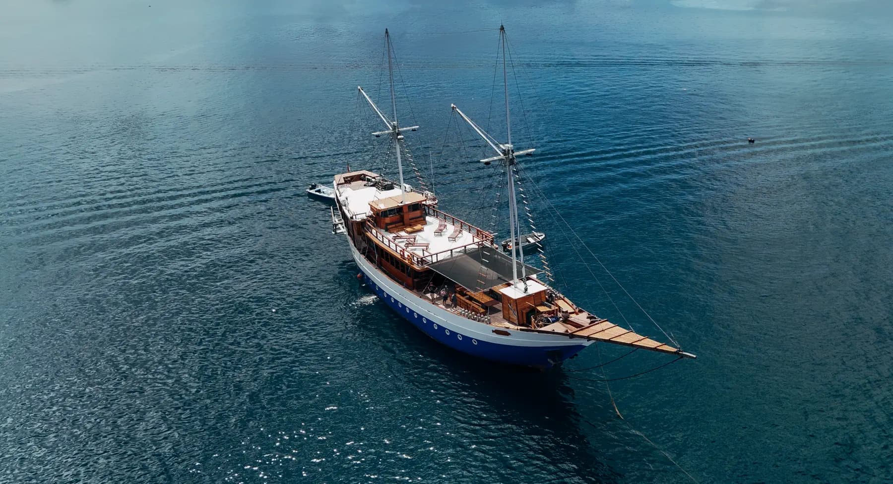 Aerial view of the Hatiku, a two-masted wooden sailing yacht, anchored on calm blue water with a gangway extended