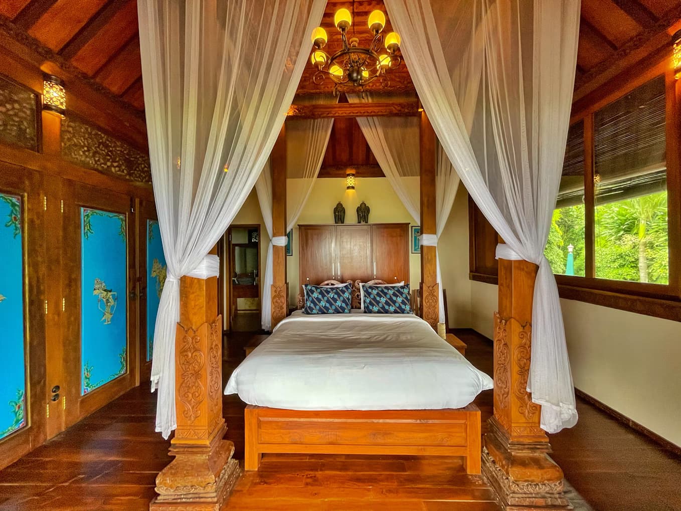Bedroom with wooden four-poster canopy bed draped in white curtains, wooden doors, ceiling fan, and windows overlooking greenery